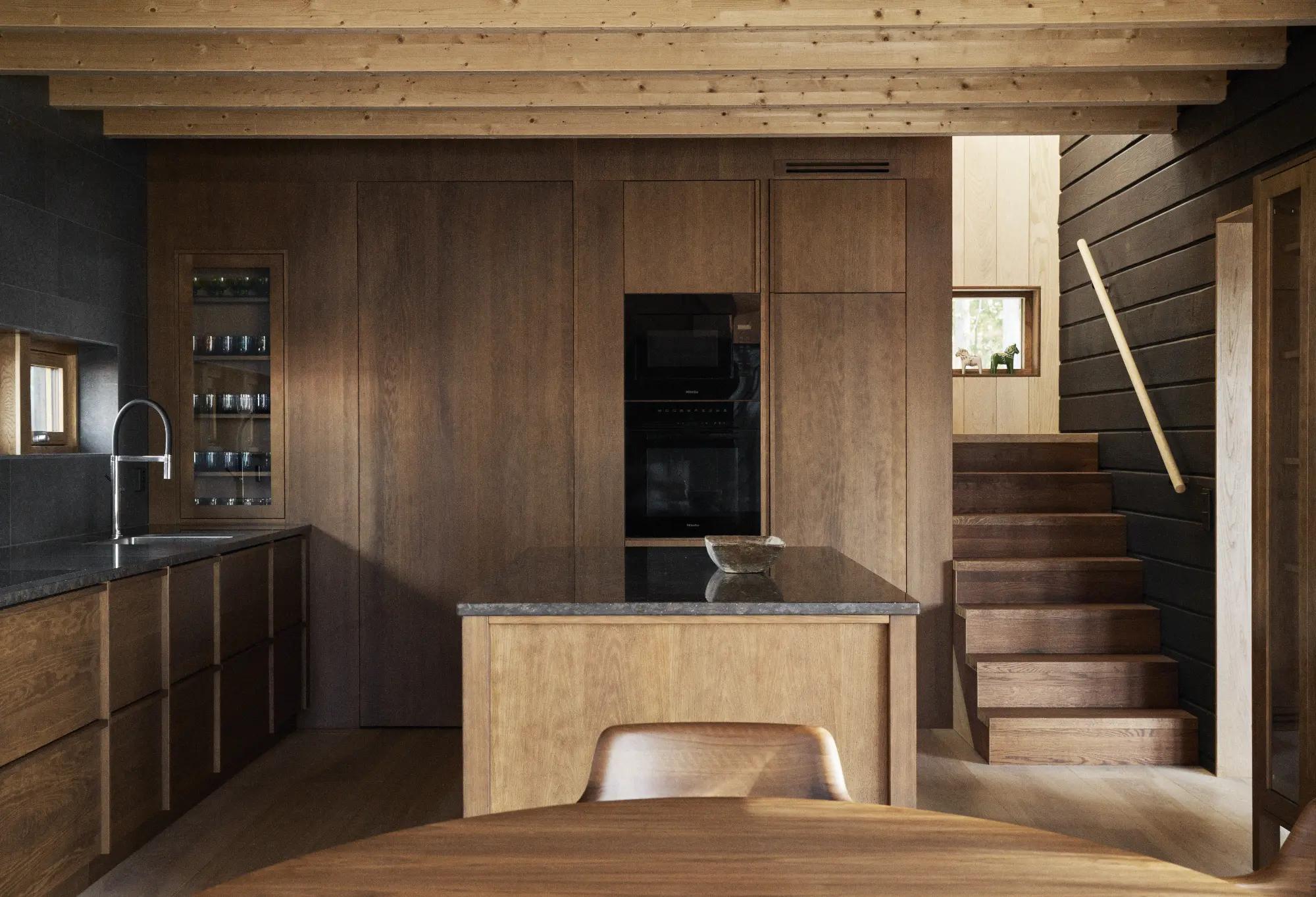 Modernt träkök med en central ö, inbyggda vitvaror och intilliggande trappa med träregelverk.