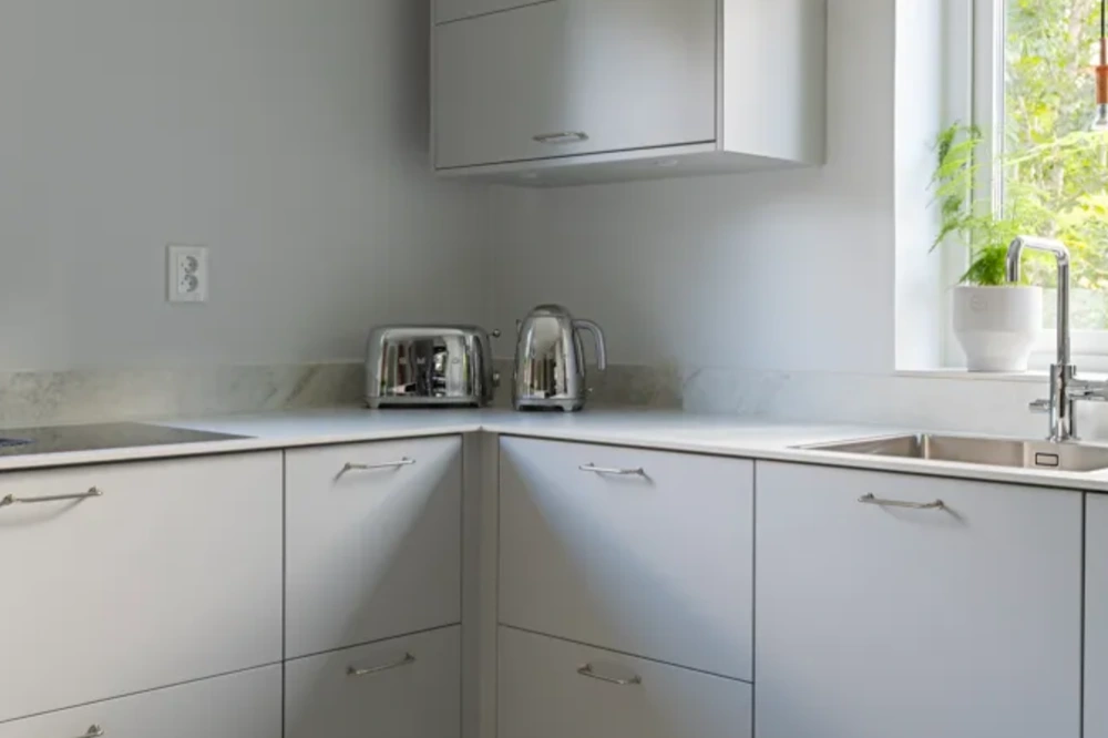 Ljust kök med grå väggar och släta, vita skåp i hörnet. På bänkskivan står två silverfärgade brödrostar. I närheten finns en diskho med en modern kran. Fönstret släpper in naturligt ljus och ramar in en kruka med en grön växt.