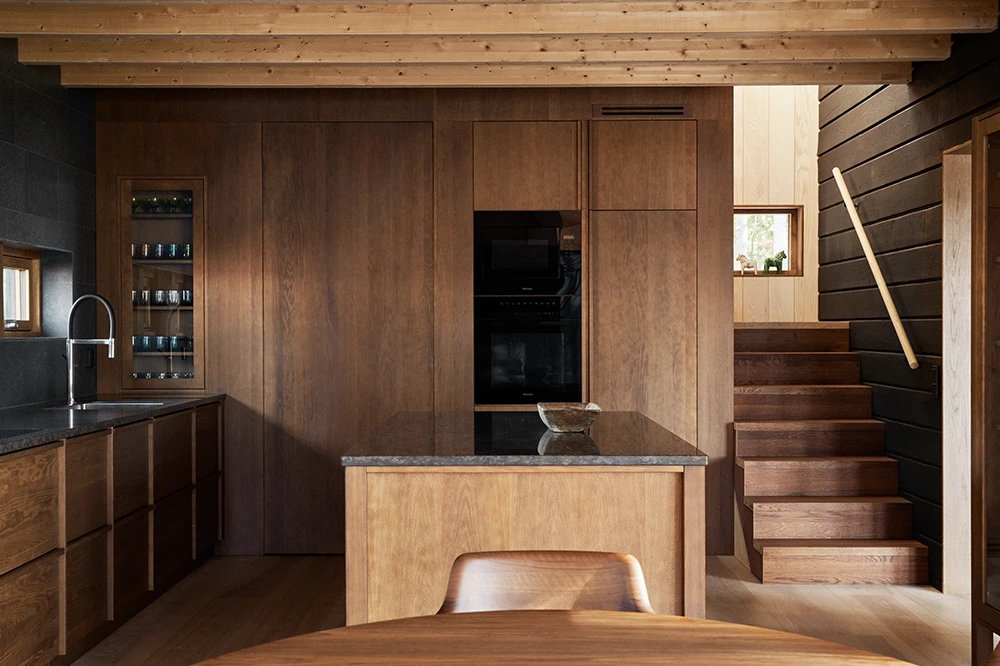 Modernt kök med skåp i mörkt trä, central köksö och trätrappa intill en vägg med panel.