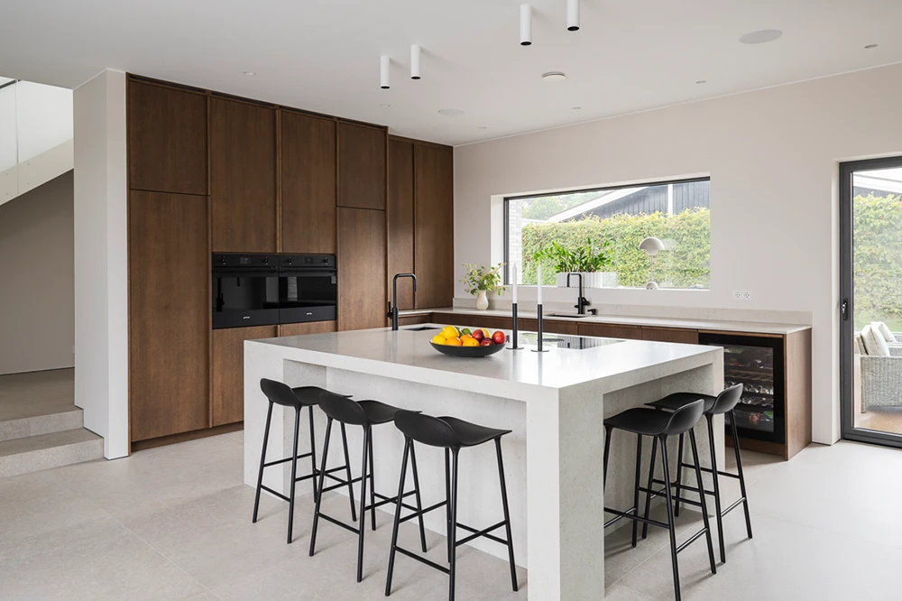 Modernt kök med träskåp, vit köksö, svarta barstolar, stort fönster och fruktskål på bänken.