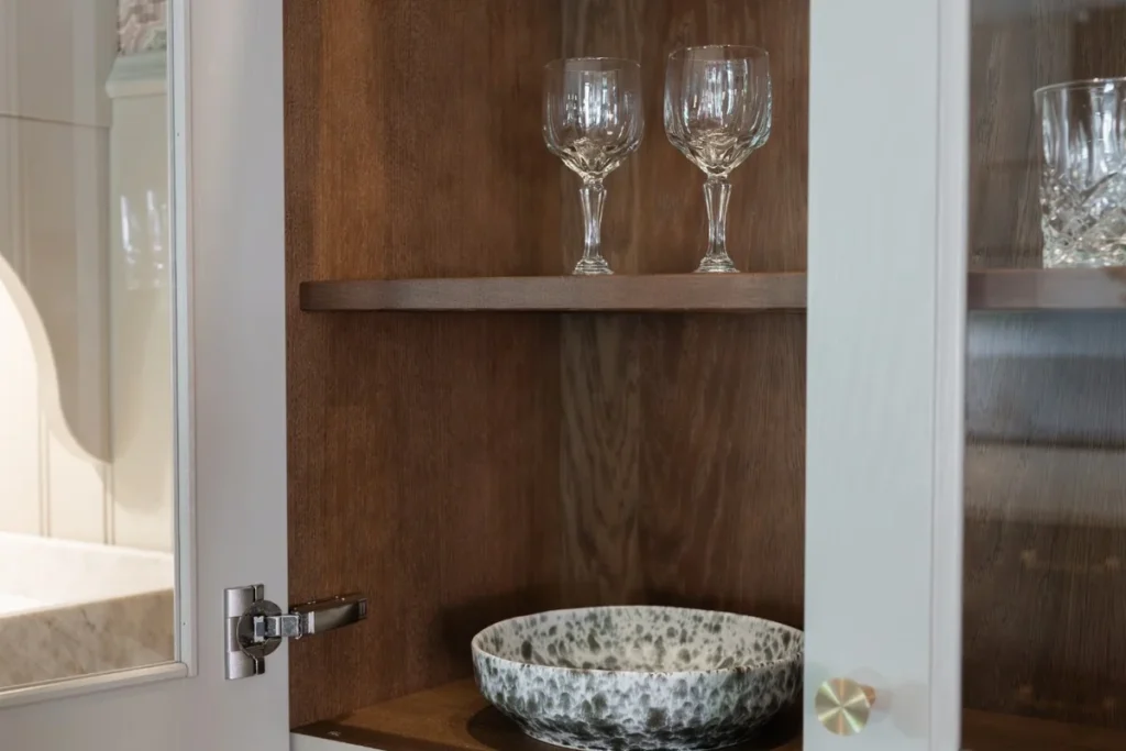 Öppnad glasad skåpdörr med insida och hyllor i brunbetsad ek, med vinglas, en skål med stänkmönster och en knopp i mässing på dörren.