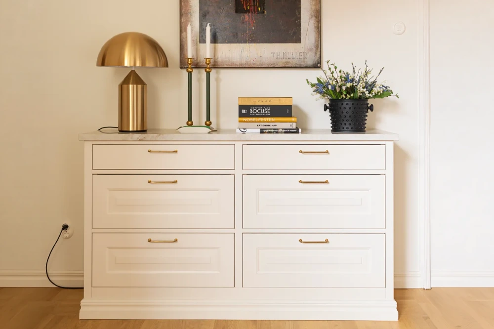 Vit kommod med sex lådor, toppad med en mörk marmorplatta. På kommodens yta står en guld färgad bordslampa, två gröna ljusstakar med ljus, och en svart kruka med blommor. Några kokböcker är staplade i raden. Väggarna är ljusa, och golvet är i trä.
