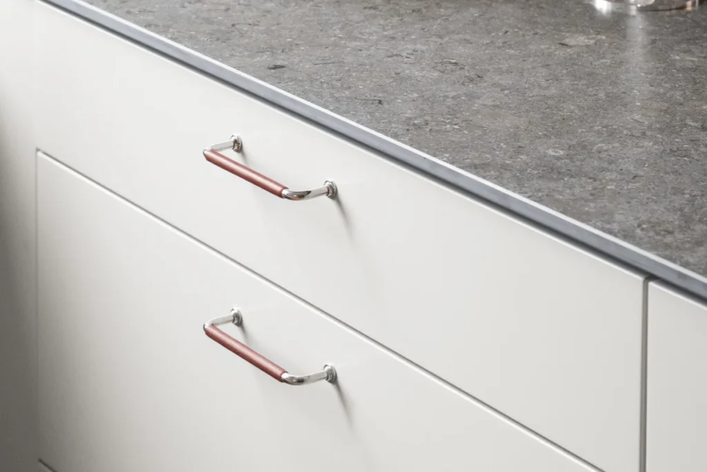 Vita, släta köksluckor med rödbruna läderhandtag i krom, under en bänkskiva i grå kompositsten. Minimalistisk Sober-design.